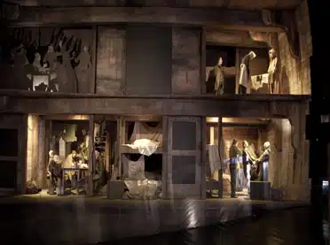 A dimly lit museum exhibit depicting a historical street scene with life-sized mannequins in various daily activities, set in detailed, period-appropriate shopfronts and homes.