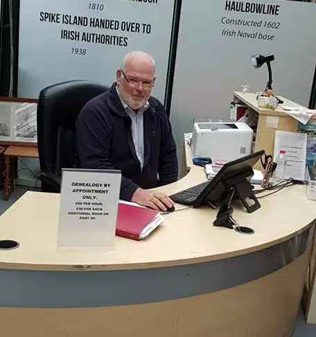 A man sitting at a desk with a computer and a sign reading "genealogy by appointment only" at an office, displaying historical posters about spike island in the background.