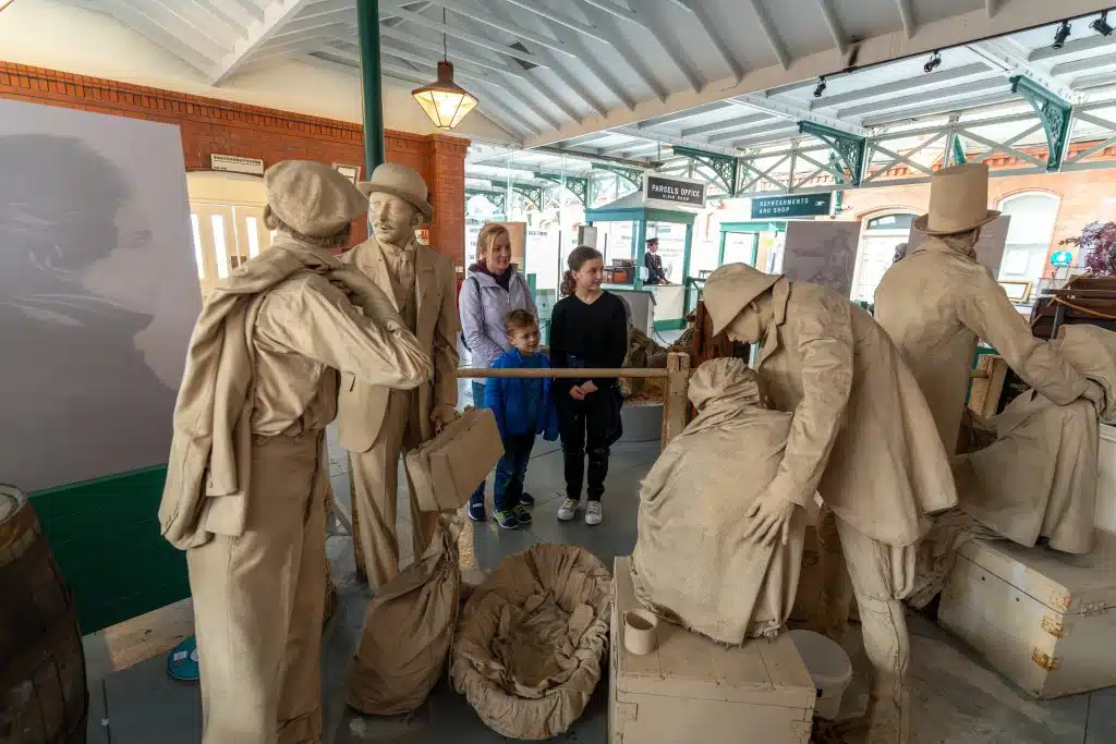 A museum display featuring life-size statues of early 20th-century travelers with luggage at a train station, interacting as a young boy and an adult observe.