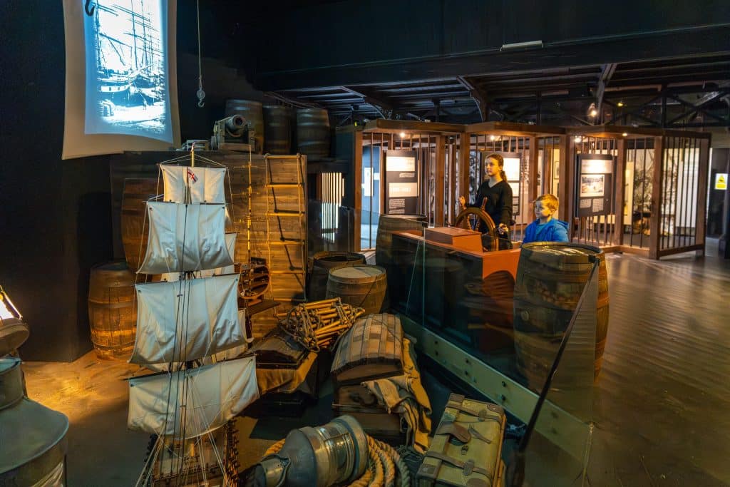 Historical exhibit with life-size mannequins. one mannequin steers a ship's wheel and another sits, surrounded by wooden barrels, crates, and nautical equipment. a ship's image is projected in the background.