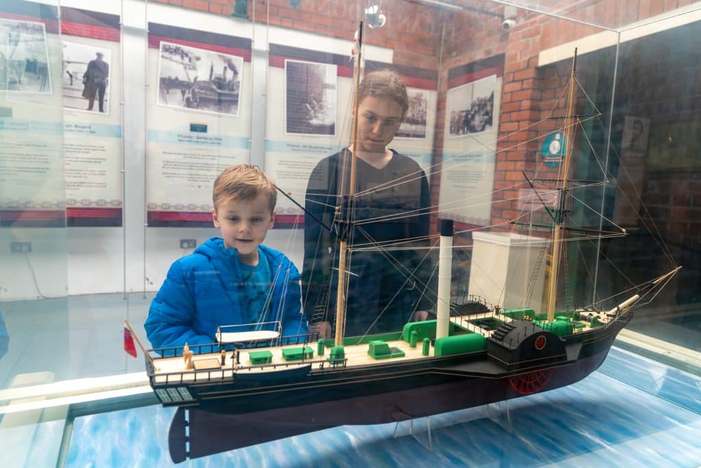 Two children, likely siblings, observing a model ship displayed inside a glass case at a museum, with informational posters visible in the background.