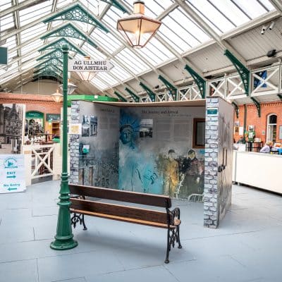 Interior of a train station with a green arched ceiling and skylights. a wooden bench faces an informational display about railway history. signs direct to shops and exits.