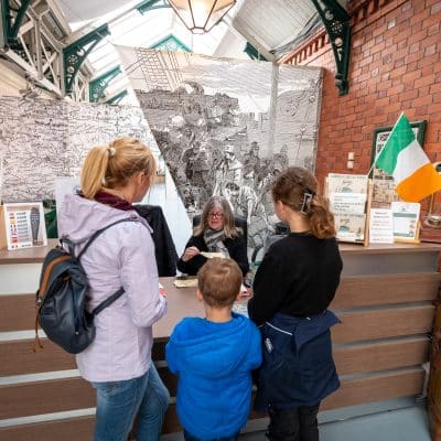 A woman educator talks to a family inside a museum with historical displays, including a large titanic illustration and an irish flag.