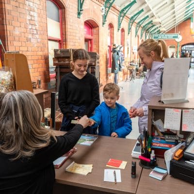 A family interacts with a staff member at a train station information desk. two girls and a young boy with their mother smile as the staff hands over tickets. the atmosphere is warm and lively.