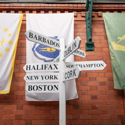 Three vertical banners hanging on a brick wall displaying maritime flags of barbados, a route map between halifax, new york, boston, and dublin, southhampton, cork, and a golden harp on a green background.