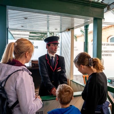 A railway station clerk dressed in a uniform interacts with a woman and two children at a ticket booth inside a museum exhibit showcasing historical timelines.