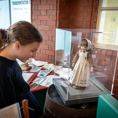 A young girl with dark hair is examining a historical doll displayed inside a transparent protective case. the exhibit is in a room with brick walls, illuminated by natural light.