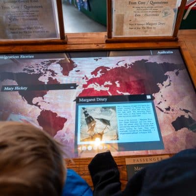 Interactive museum display showing a map-based digital screen with emigration stories to australia, viewed over the shoulder of a visitor. text and images detail individual histories.