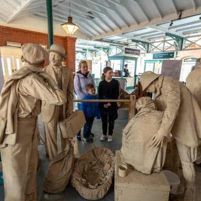 A museum display depicting life-sized statues of vintage railway passengers and staff in an old station setting, interacting around suitcases, with two real visitors observing.