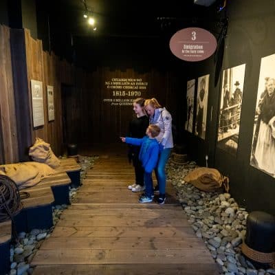 Two children explore a museum exhibit about emigration, with displays that include a wooden boat, ropes, and informational posters on the walls.
