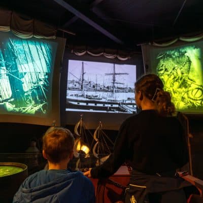 A woman and a young boy sit facing large screens displaying historical ship images, in a dark, immersive exhibit room with ship models on the table in front of them.