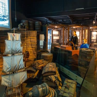 An exhibit displaying maritime artifacts, including crates, barrels, and ship equipment, with two mannequins, one steering a wheel. a large vintage ship photo and video are projected in the background.