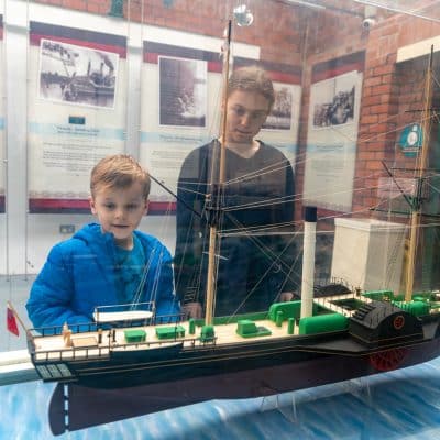 Two children, a young boy in a blue jacket and an older boy in a striped shirt, look intently at a model ship displayed in a glass case at a museum.