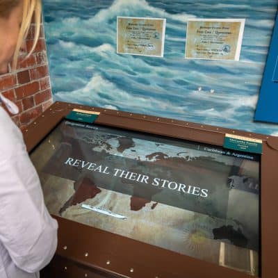 A visitor interacts with an interactive museum exhibit titled "reveal their stories" featuring a digital touchscreen showcasing historical content, set against a backdrop of ocean-themed murals.