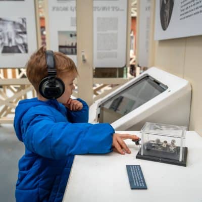 A young child wearing headphones and a blue jacket touches a display at a museum exhibit, engrossed in learning from an interactive screen.