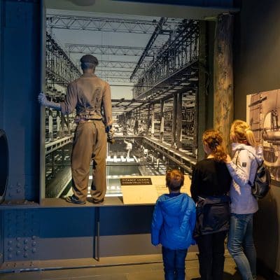 Visitors, including children and adults, observe a large photographic exhibit featuring an historical industrial scene with a life-size figure of a worker in the foreground.