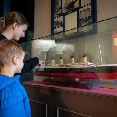 A woman and a young boy are looking at a model ship displayed inside a glass case in a museum. the woman is pointing something out to the boy as they study the model intently.