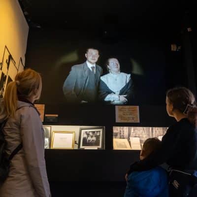 Visitors at a museum exhibition viewing a black and white projected image of a smiling man and woman, surrounded by informational displays and artifacts related to the titanic.