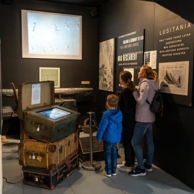 A family observes a display at a museum about the lusitania, featuring old luggage, documents, and a background video. the exhibit includes detailed informational boards.