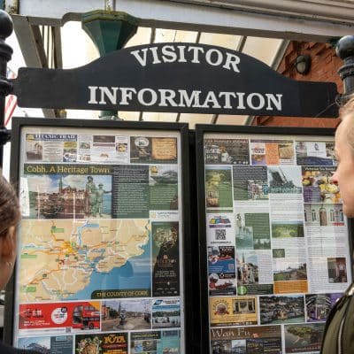 Two women look at a visitor information board with brochures and maps under a sign that reads "visitor information" in a town setting.