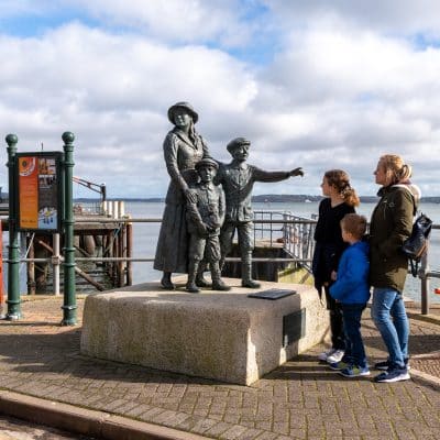 A statue of a family with two young children and a mother pointing, situated at a pier. real visitors, a family of four, interact with the monument, overlooking the water on a cloudy day.