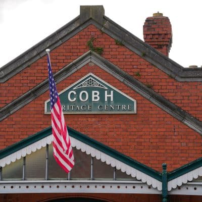 The façade of the cobh heritage centre in ireland, featuring a red brick wall with the name embedded in white tiles and an american flag hanging prominently in front.