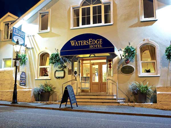 Nighttime view of the watersedge hotel entrance with a blue canopy, illuminated by warm lights, featuring hanging plants and a welcoming sign by the door.
