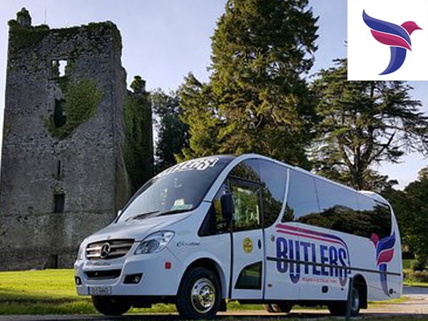 A butler's bus parked in front of an ancient stone tower surrounded by lush green trees under a clear sky.