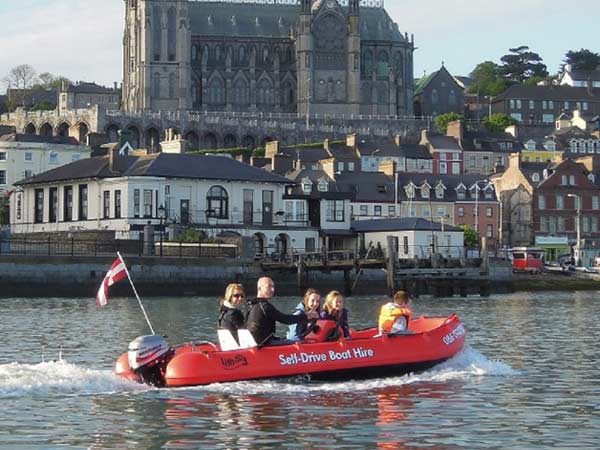 A group of four people enjoying a ride in a red self-drive hire boat on a river, with a large cathedral and hillside buildings in the background.