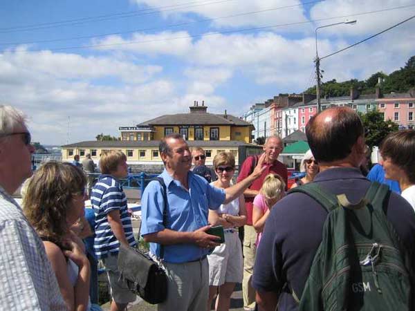 A group of tourists on a sunny day, listening to a guide in a town with colorful buildings and clear blue sky in the background.