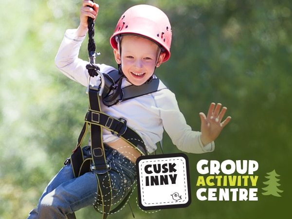 A joyful child wearing a safety helmet and harness enjoys a zip line at an outdoor group activity center, surrounded by lush greenery.