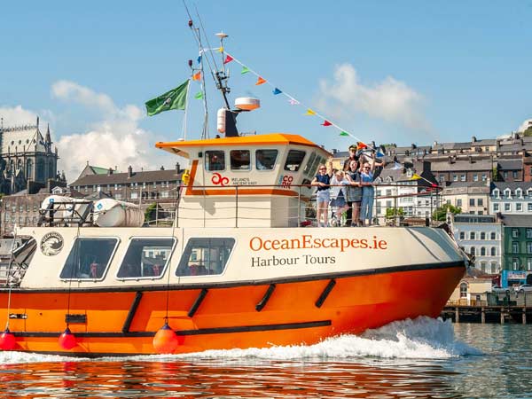 A bright orange oceanescapes.ie tour boat with passengers on board travels through a harbor with buildings visible in the background on a sunny day.
