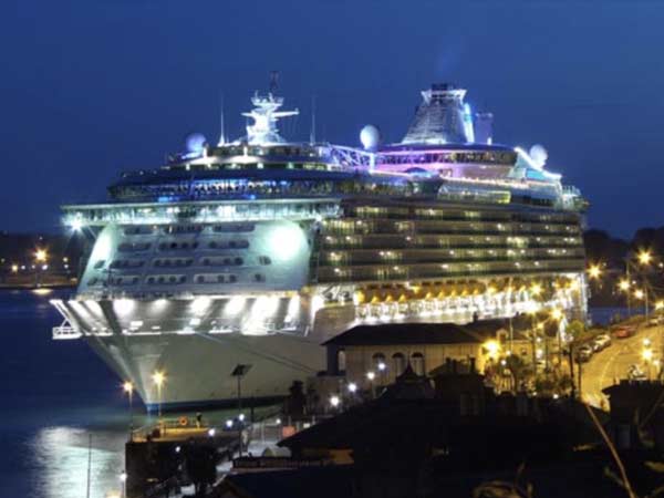 A large cruise ship illuminated at night, docked at a busy port with glowing streetlights and a deep blue twilight sky in the background.