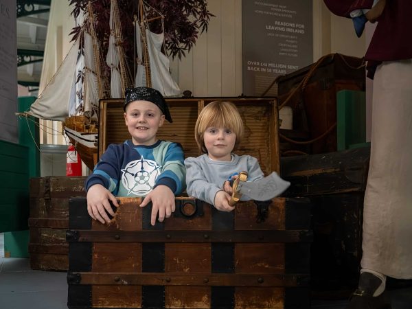 Two young children, a boy and a girl, smiling inside a vintage wooden chest at a museum exhibit, with dried plants and historical displays in the background.